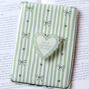 May include: A clear, protective case for an e-reader, featuring a green and white striped pattern with bows and hearts. A heart-shaped sticker with the text "born to read, forced to work" is attached.