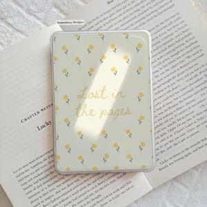 May include: A rectangular phone case featuring a floral design of yellow flowers on a cream backdrop. The text "Lost in the pages" is in the centre. The case rests on an open book.