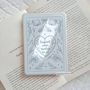 May include: A clear rectangular acrylic block featuring a blue line art illustration of a heart with the text "happiest when reading" surrounded by floral motifs, open books, and a bow, resting on an open book with visible printed text.