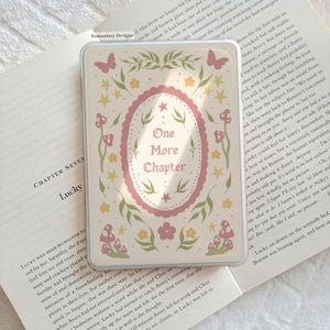 May include: A rectangular e-reader case featuring a cream background with a central pink scalloped oval frame containing the text One More Chapter. The design includes green leaves, yellow flowers, pink mushrooms, and small pink butterflies.