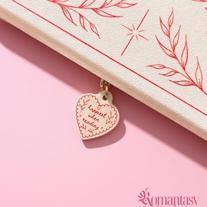 May include: A cream-coloured book cover featuring red line art of leaves and stars. A heart-shaped charm hangs from the spine, inscribed with the text 'happiest when reading' in red, surrounded by a decorative scalloped border and leaf motifs.