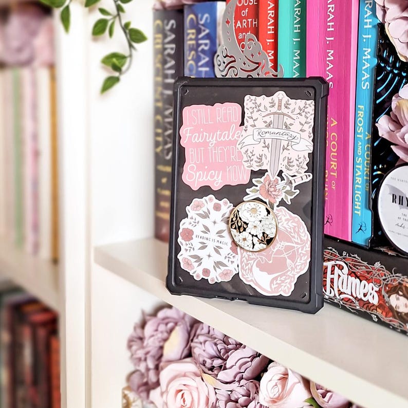 May include: A black e-reader case adorned with pink floral stickers and a pop socket. The stickers include text that reads "I STILL READ Fairytales BUT THEY'RE Spicy NOW" and "Reading is Magic". Bookshelves are in the background.