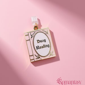 May include: A gold-toned metal enamel charm shaped like a book with a light pink cover and a white oval center. The text Busy Reading is printed in black in the center. The charm features a small clear plastic attachment at the top loop.