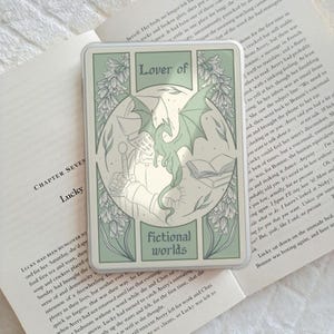 May include: A rectangular sticker with a sage green and white colour palette featuring a central illustration of a dragon, a knight in armour, and an open book. The text reads Lover of fictional worlds in a decorative serif font style.