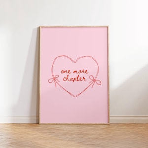 May include: A framed pink art print with a red heart outline and the words "one more chapter." The heart has two bows and the print is in a light wood frame. The artwork is leaning against a white wall on a wooden floor.