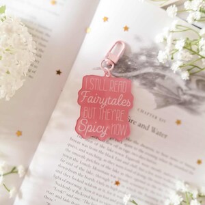 May include: A pink keychain with the text "I STILL READ Fairytales BUT THEY'RE Spicy NOW." The keychain is attached to a pink clasp. The background includes a book and white flowers.