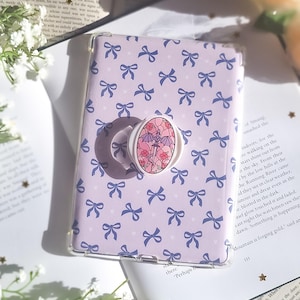May include: A lilac phone case featuring a pattern of blue bows and tiny hearts. A white pop socket with a floral design is attached. The case rests on an open book.