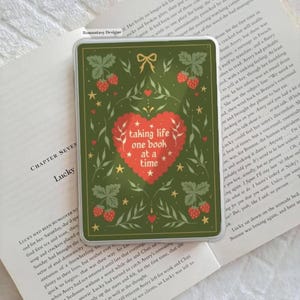 Peut inclure: Une coque de téléphone rectangulaire verte avec un motif de cœur rouge indiquant "taking life one book at a time". La coque est ornée de fraises, de feuilles et d'étoiles. La coque est posée sur un livre ouvert.