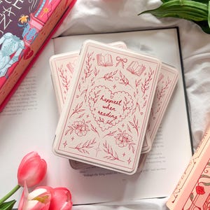 Könnte beinhalten: Eine cremefarbene E-Reader-Hülle mit einer roten Strichzeichnung eines Herzens, floralen Ranken, offenen Büchern und Schleifen. Der Text "happiest when reading" ist mittig im Herzen platziert. Die Hülle liegt auf einem aufgeschlagenen Buch.