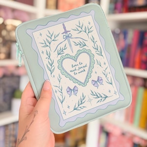 May include: A sage green rectangular tablet or book sleeve with a zip closure. The front features a decorative frame with a central heart containing the text "born to read, forced to work," surrounded by illustrations of a sword, bows, and leaves.