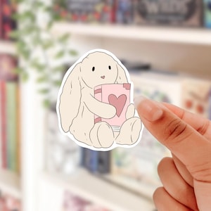 May include: A white bunny with long ears holding a pink book with a heart on the cover. The book has the words "Love Story" on the cover.
