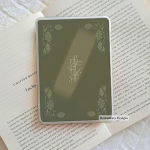 May include: A rectangular e-reader case in a muted olive green shade, featuring a delicate white line art design of a central sword entwined with vines, surrounded by a border of various leaf illustrations at each of the four corners.