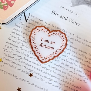 May include: A heart-shaped sticker with a brown lace-like border and the text I am an Autumn printed in the centre in a dark brown serif font. The sticker is placed on a page of a book with visible printed text and small gold star accents.