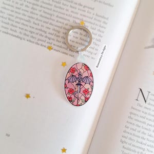 May include: Oval keychain with a dragon and sword design, framed by pink flowers, in a stained-glass style. The dragon has purple wings. The keychain has a silver ring and is attached to a clear bead.
