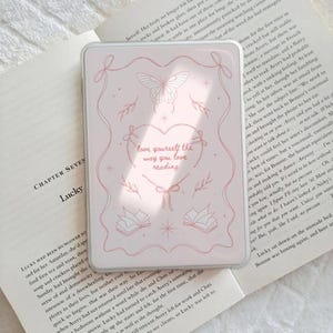 Könnte beinhalten: Eine transparente Handyhülle mit rosa Design. Sie zeigt einen Schmetterling, Herzen, Schleifen und den Text "love yourself the way you love reading". Die Hülle liegt auf einem offenen Buch.