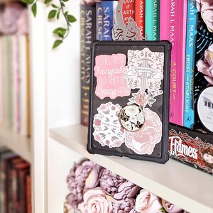 May include: A black case with a variety of stickers featuring floral designs, a sword, and text that reads "I still read Fairytales but they're Spicy Now" and "Romantasy".