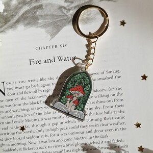 Peut inclure: Un porte-clés en acrylique transparent avec un anneau et une chaîne dorés. Le porte-clés présente un design fantaisiste de champignons rouges et blancs à l'intérieur d'une fenêtre arquée teintée de vert, sur un livre ouvert. Le texte "Fire and Water" est visible en arrière-plan.