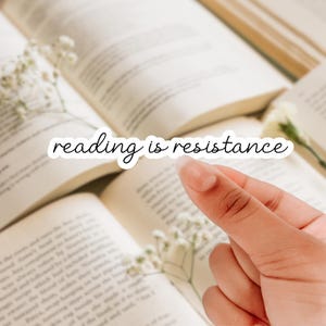 May include: A sticker with the words "reading is resistance" in a cursive font is placed over an open book. The background features more open books and delicate white flowers.