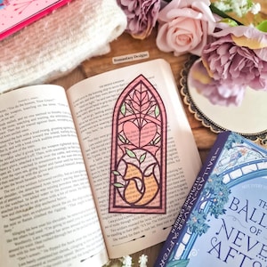 May include: A pink stained glass bookmark with a heart and fox design, resting on an open book. The bookmark has a decorative border and is surrounded by flowers and a book titled "The Ballad of Never After".