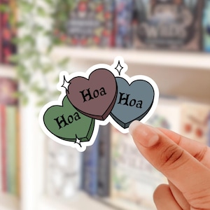 May include: Three heart-shaped stickers in green, brown, and blue, each with the word "Hoa" in black. The stickers have a white outline and star accents. The stickers are held by a person's hand.