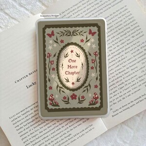 May include: A rectangular phone case with a whimsical design. The case features an oval with the text "One More Chapter" surrounded by floral and mushroom illustrations in shades of green, red, and white.