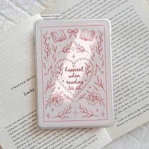 May include: A white and pink phone case with the text "happiest when reading" in a heart-shaped design. The case features floral and botanical illustrations. The case is resting on an open book.