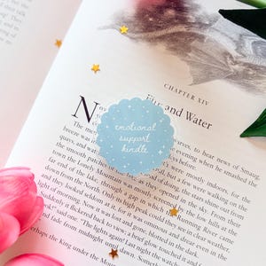 May include: A circular light blue sticker with a scalloped edge featuring white star accents and the handwritten text "emotional support kindle" in white script, placed on an open book page with printed text and small gold star confetti.