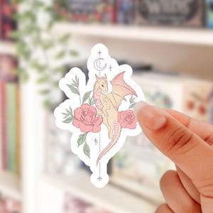 May include: A sticker featuring a light yellow dragon with pink wings, surrounded by pink roses and green leaves. The design includes a crescent moon and stars. The sticker is held by a hand.