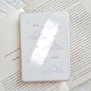 May include: A clear phone case featuring a light blue design with the words "head in the clouds" and illustrations of clouds. The case is shown on top of an open book.