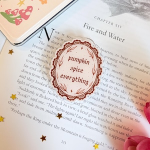 May include: An oval-shaped acrylic pin featuring a brown lace-style border and the text "pumpkin spice everything" in brown lettering. The design includes small leaf and star accents, centered on a page of a book titled Chapter XIV Fire and Water.