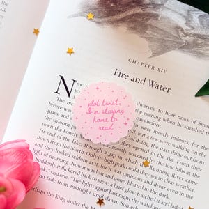 May include: A circular, scalloped-edge sticker with a light pink background and small star accents. The sticker features the dark pink text 'plot twist, I'm staying home to read' centred on the surface, placed on an open book page with text.