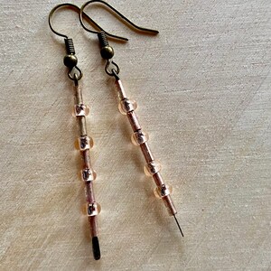 Pu&ograve; includere: Un paio di orecchini lunghi e delicati con un gancio in bronzo e una catena di piccole perle rosa traslucide.