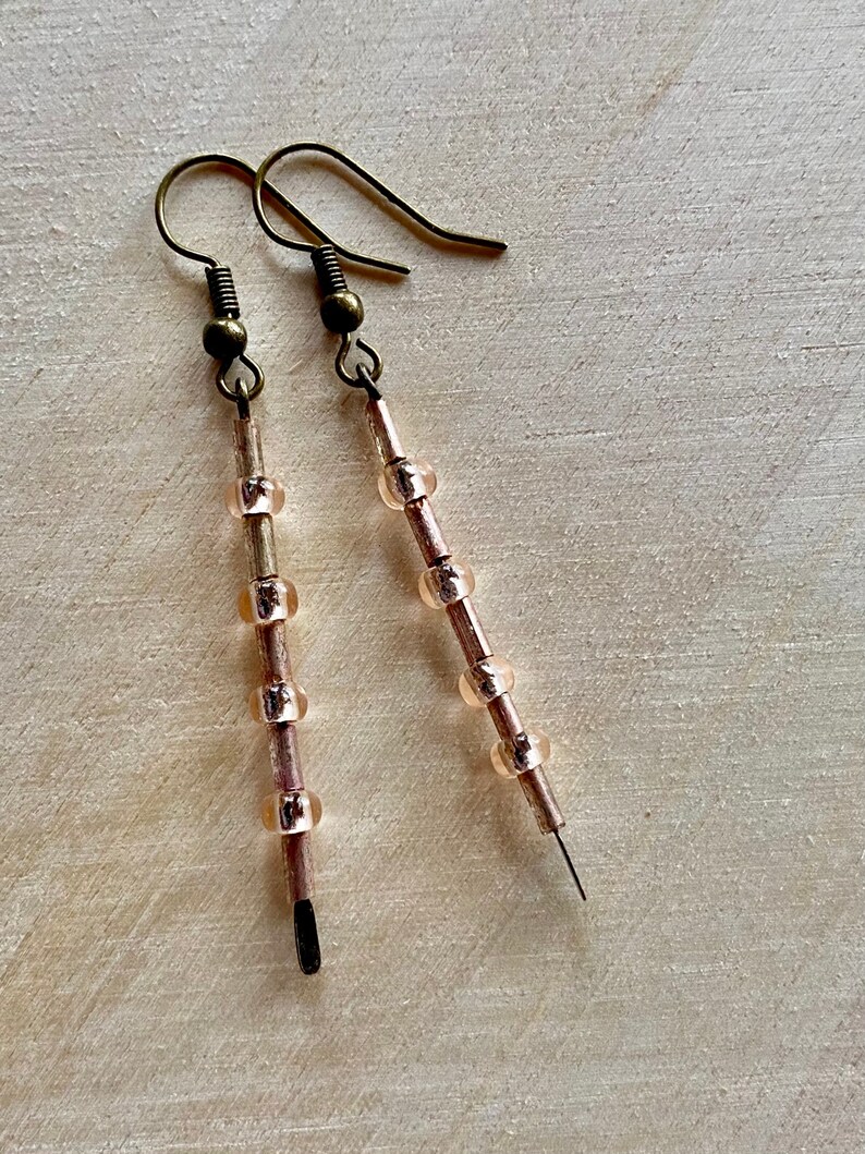 Pu&ograve; includere: Un paio di orecchini lunghi e delicati con un gancio in bronzo e una catena di piccole perle rosa traslucide.