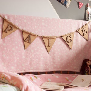 May include: A wooden banner with the name "Paige" spelled out in individual letters. The banner is hanging above a pink cot with a pink and white blanket and a pink and white sheet.