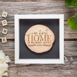 May include: A framed wooden slice with the text "Our First Home" and the address "21 Mulberry Avenue 2021" engraved on it. The frame is white, and the background is black. The wooden slice is circular and has visible wood grain.