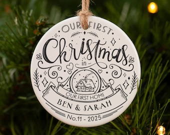 May include: A white ceramic Christmas ornament with black lettering and a twine hanger. The ornament reads "Our First Christmas in Our First Home" with the names "Ben & Sarah" and the date "No.11-2025". The ornament is hanging on a Christmas tree.