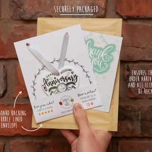 May include: A white ceramic heart-shaped ornament with the text "Happy Wedding Anniversary" and a small number "1" on it. The ornament is packaged in a brown envelope with a white card that says "Thank You". The package is secured with a cardboard backing and bubble-lined envelope.