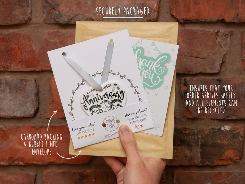 May include: A white ceramic heart-shaped ornament with the text "Happy Wedding Anniversary" and a small number "1" on it. The ornament is packaged in a brown envelope with a white card that says "Thank You". The package is secured with a cardboard backing and bubble-lined envelope.