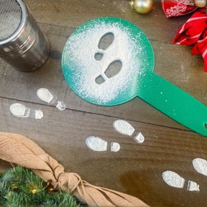 Peut inclure: Un pochoir vert en forme d'empreintes de pas, utilisé pour saupoudrer du sucre glace et créer des empreintes sur une surface en bois. Un shaker en acier inoxydable et des décorations de Noël sont également présents.