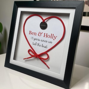 May include: Framed artwork featuring a red shoelace heart on a white background. The text "Ben & Holly" and "13 years since we tied the knot" are printed in red. The frame is black.