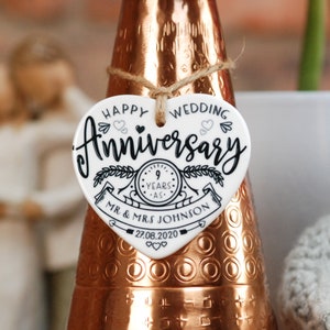 May include: White ceramic heart-shaped ornament with black text that reads "Happy Wedding Anniversary 9 Years - Mr. & Mrs. Johnson 27.08.2020". The ornament has a black and white illustration of a clock with a ribbon and leaves.