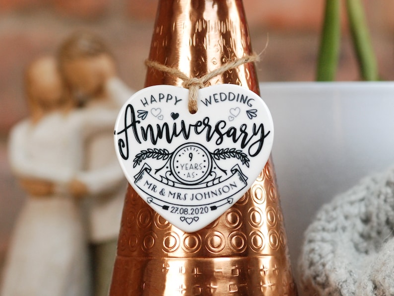 May include: White ceramic heart-shaped ornament with black text that reads "Happy Wedding Anniversary 9 Years - Mr. & Mrs. Johnson 27.08.2020". The ornament has a black and white illustration of a clock with a ribbon and leaves.