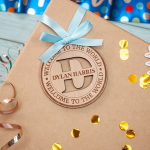 A brown gift box with a blue ribbon and a wooden tag that says "Welcome to the World Dylan Harris Welcome to the World". The tag has a large letter "D" in the centre.