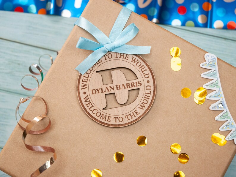 A brown gift box with a blue ribbon and a wooden tag that says "Welcome to the World Dylan Harris Welcome to the World". The tag has a large letter "D" in the centre.