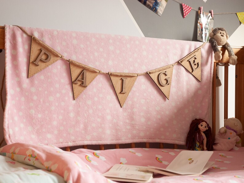 May include: A wooden banner with the name "Paige" spelled out in individual letters. The banner is hanging above a pink cot with a pink and white blanket and a pink and white sheet.