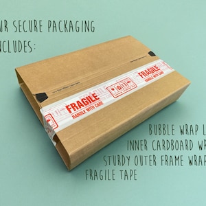 May include: A brown cardboard box with a white and red "FRAGILE" label and black tape. The box is on a light blue background. The text "OUR SECURE PACKAGING INCLUDES:" is above the box. The text "BUBBLE WRAP LAYERS", "INNER CARDBOARD WRAP", "STURDY OUTER FRAME WRAP", and "FRAGILE TAPE" is below the box.