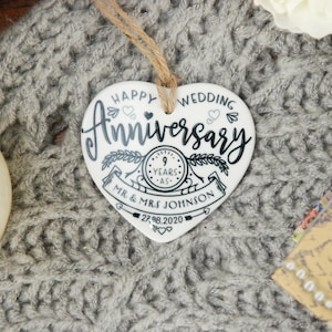 May include: White ceramic heart-shaped ornament with black text that reads "Happy Wedding Anniversary" with a clock graphic that says "9 Years" and "Mr. & Mrs. Johnson" with the date "27.8.2020".