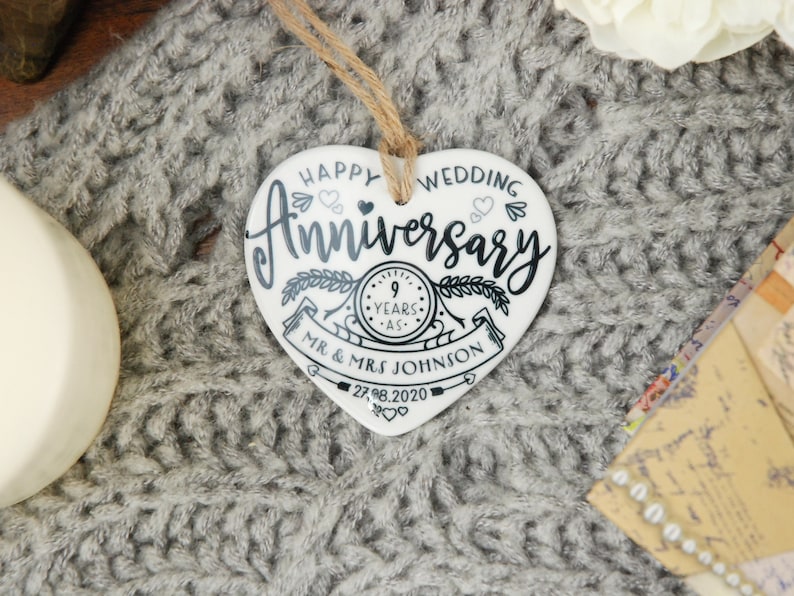 May include: White ceramic heart-shaped ornament with black text that reads "Happy Wedding Anniversary" with a clock graphic that says "9 Years" and "Mr. & Mrs. Johnson" with the date "27.8.2020".