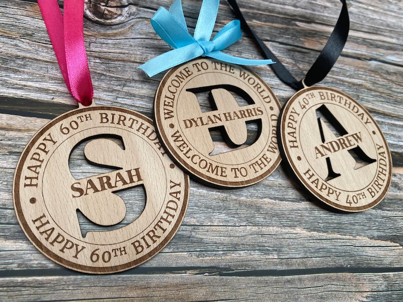 May include: Three wooden round decorations with a ribbon attached. Each decoration has a different name and age inscribed on it. The first decoration says "Happy 60th Birthday Sarah". The second decoration says "Welcome to the World Dylan Harris". The third decoration says "Happy 40th Birthday Andrew".