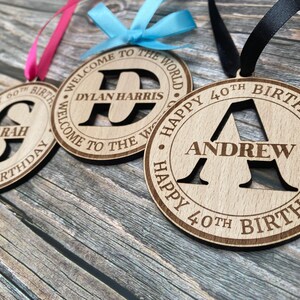 Three wooden decorations with a personalised initial and name. The first decoration is pink and says "Happy 60th Birthday Sarah". The second decoration is blue and says "Welcome to the World Dylan Harris". The third decoration is black and says "Happy 40th Birthday Andrew".
